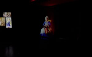 Holographic figure of a seated woman in historical costume on a dark stage with small screens at left.