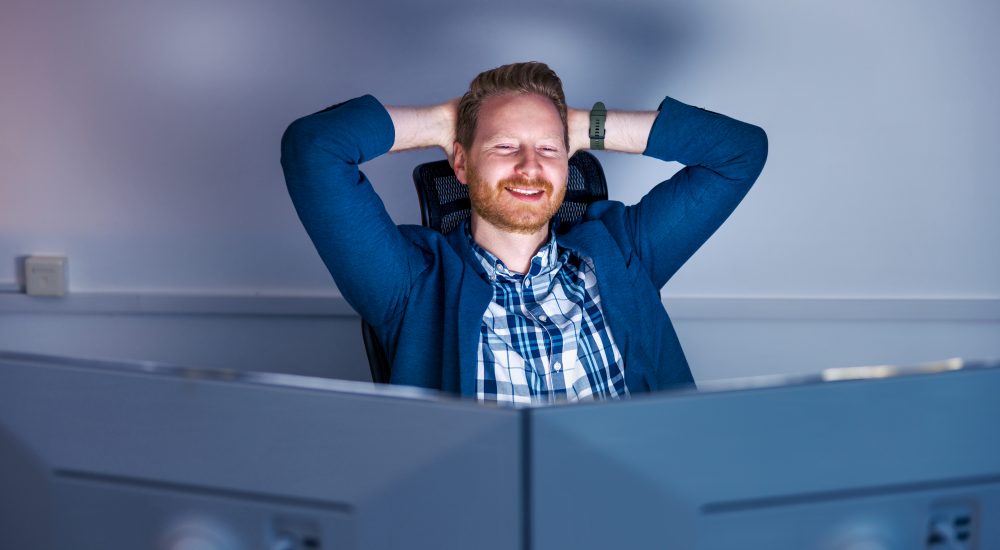 Casual businessman working late in an office Confident successful businessman sitting at his desk using desktop computer while working late in an office, smiling and looking towards computer screen